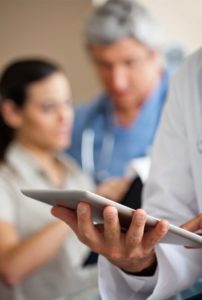 Doctor looking at iPad with nurse and patient in background| TeleMed Inc.